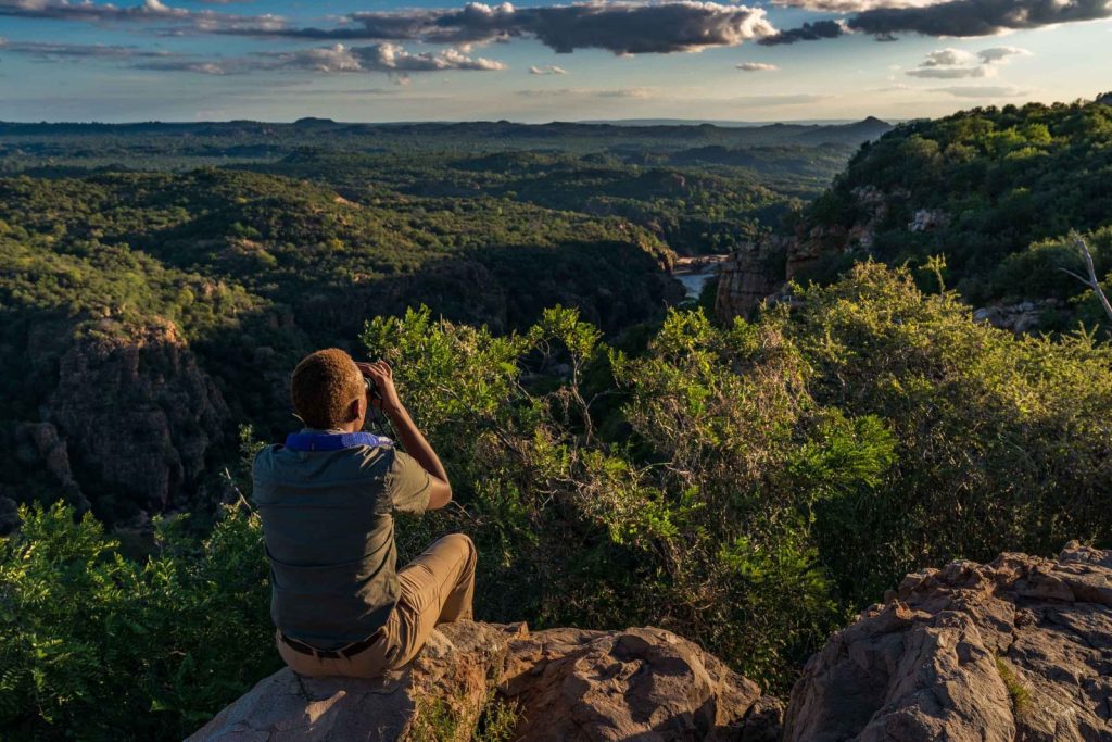 A safari guide sat on a rock with binoculars overlooking green tree-covered mountains